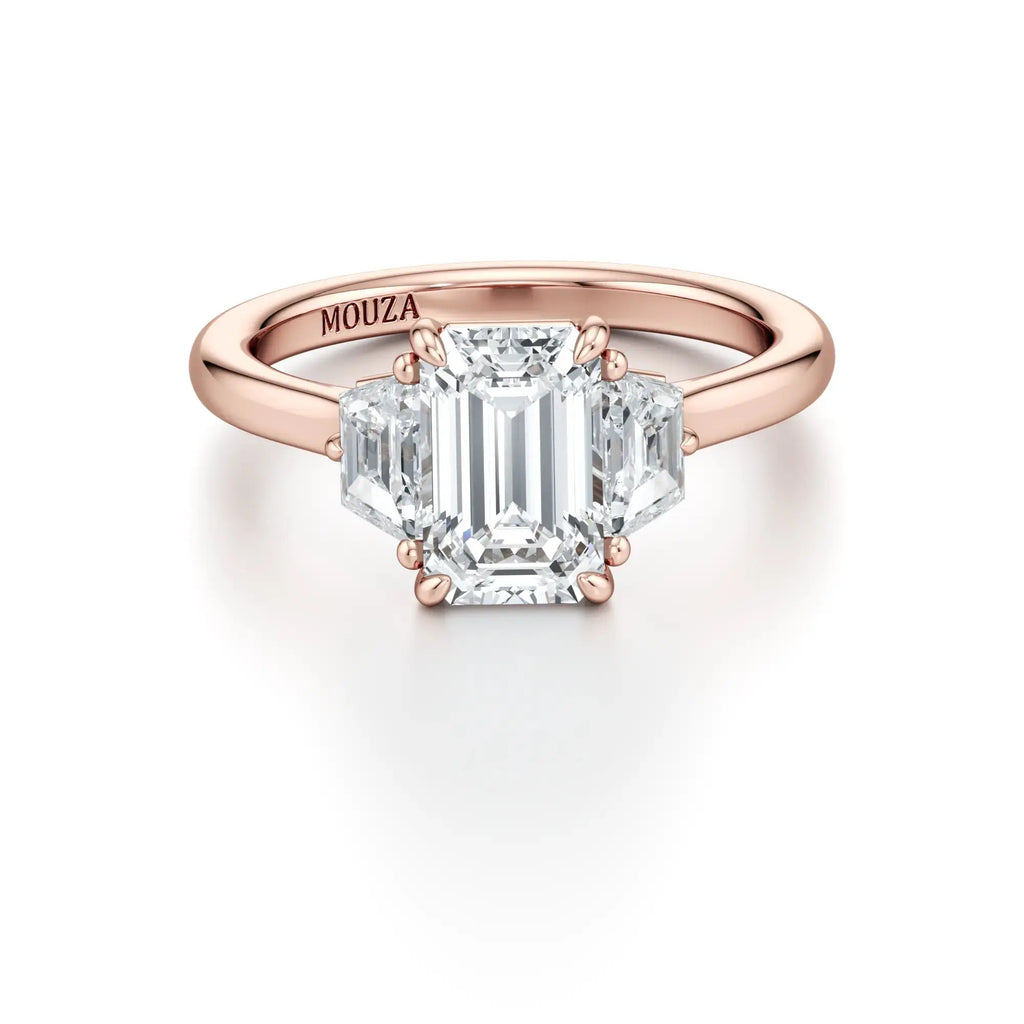 A sleek engagement ring featuring a central emerald-cut diamond flanked by two trapezoid-cut side stones, set in a rose gold band with the brand name 'Mouza' visible on the side.