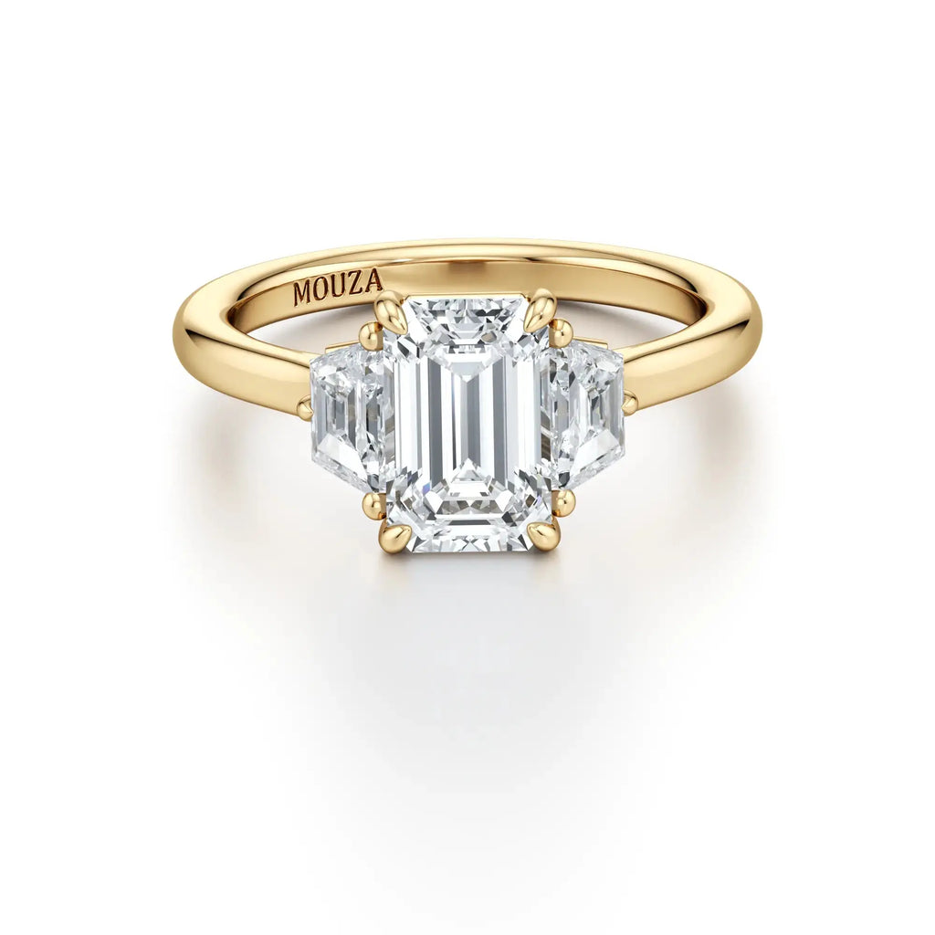 A sleek engagement ring featuring a central emerald-cut diamond flanked by two trapezoid-cut side stones, set in a gold band with the brand name 'Mouza' visible on the side.