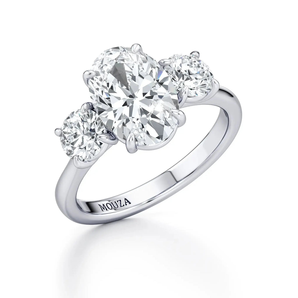 A classic three-stone engagement ring with a central oval diamond, having the brand name 'Mouza' on it.
