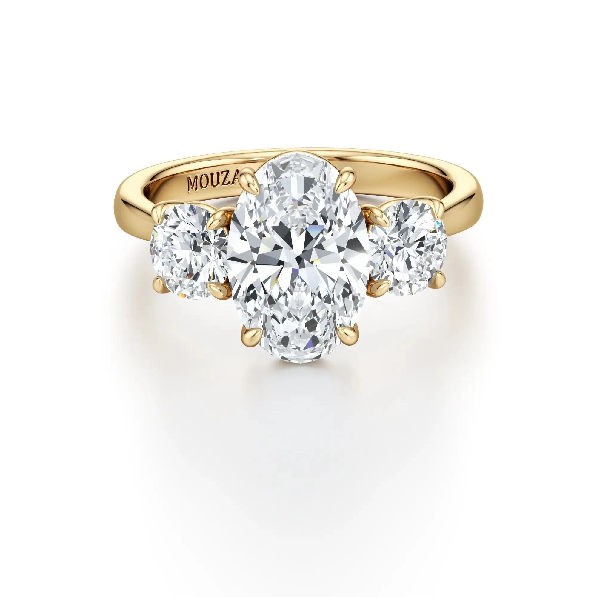 A classic three-stone engagement ring with round side stones and a central oval diamond, with a gold band having the brand name 'Mouza' on it.