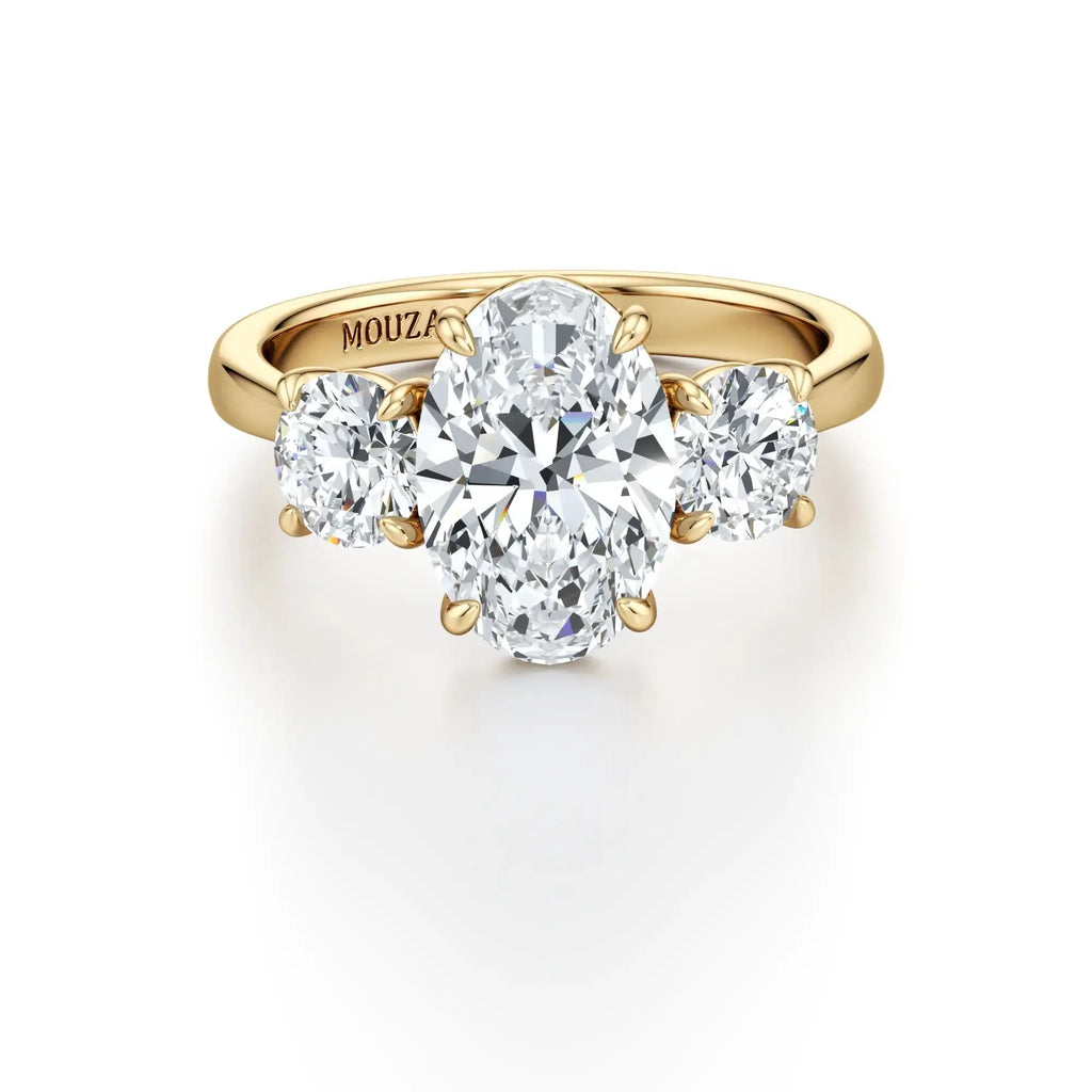 A classic three-stone engagement ring with round side stones and a central oval diamond, with a gold band having the brand name 'Mouza' on it.