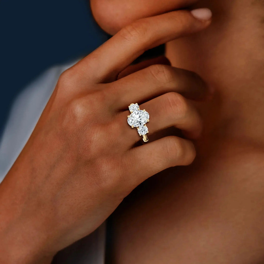 A classic trilogy engagement ring with round side stones and a central oval diamond, with a gold band having the brand name 'Mouza' on it.